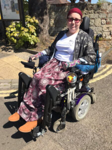 A white woman, wearing tinted glasses and a red head-wrap, smiling at the camera sitting in her powerchair.