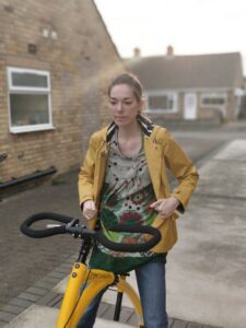 A white woman with brown hair, wearing a yellow coat and using a walker.