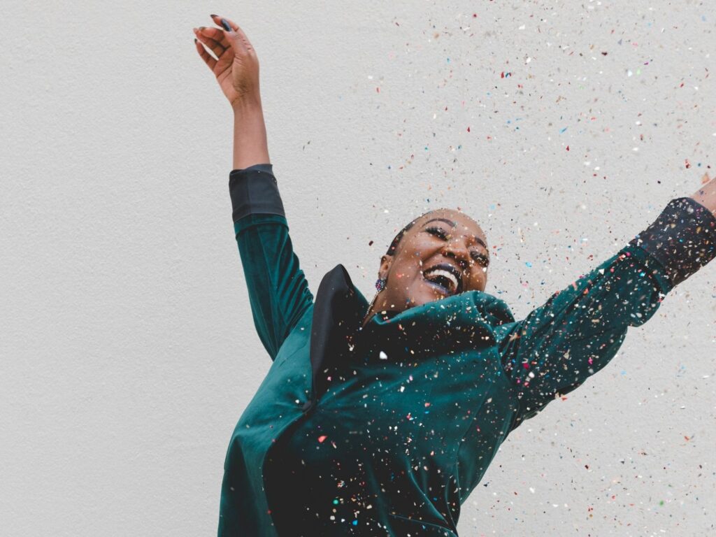Someone smiling and raising their arms joyfully. Coloured glitter is falling on them from above.