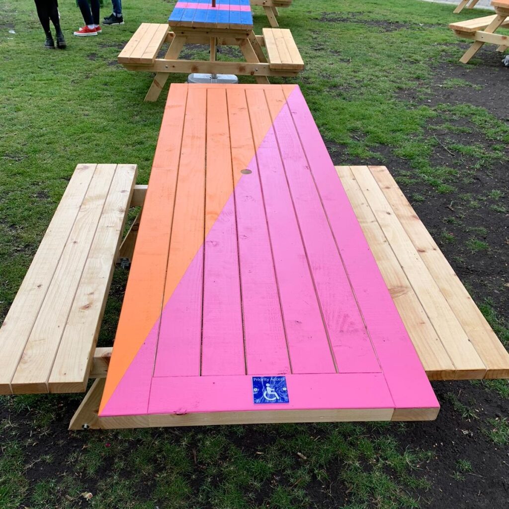 A wooden picnic table with fixed benches on either side. One end of the tabletop projects towards the camera and has a 'wheelchair symbol' on it