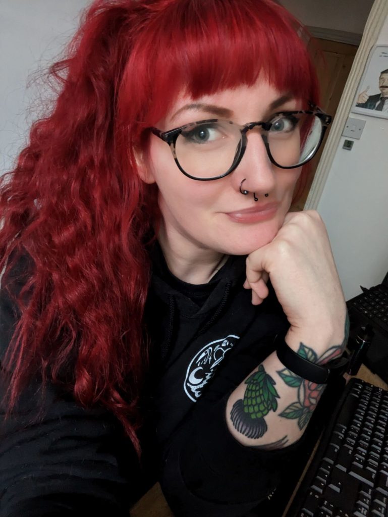 Photo of Carla with red hair tied up, thick glasses and wearing a black hoodie