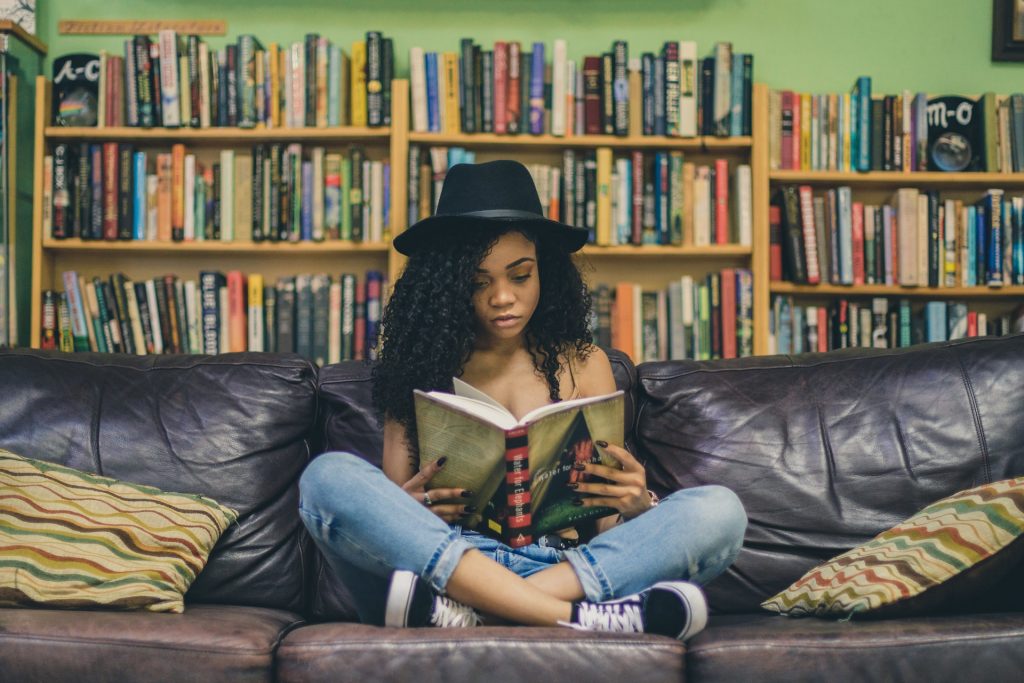 Girl sat cross legged on a sofa reading a book. Shelves of books behind her.