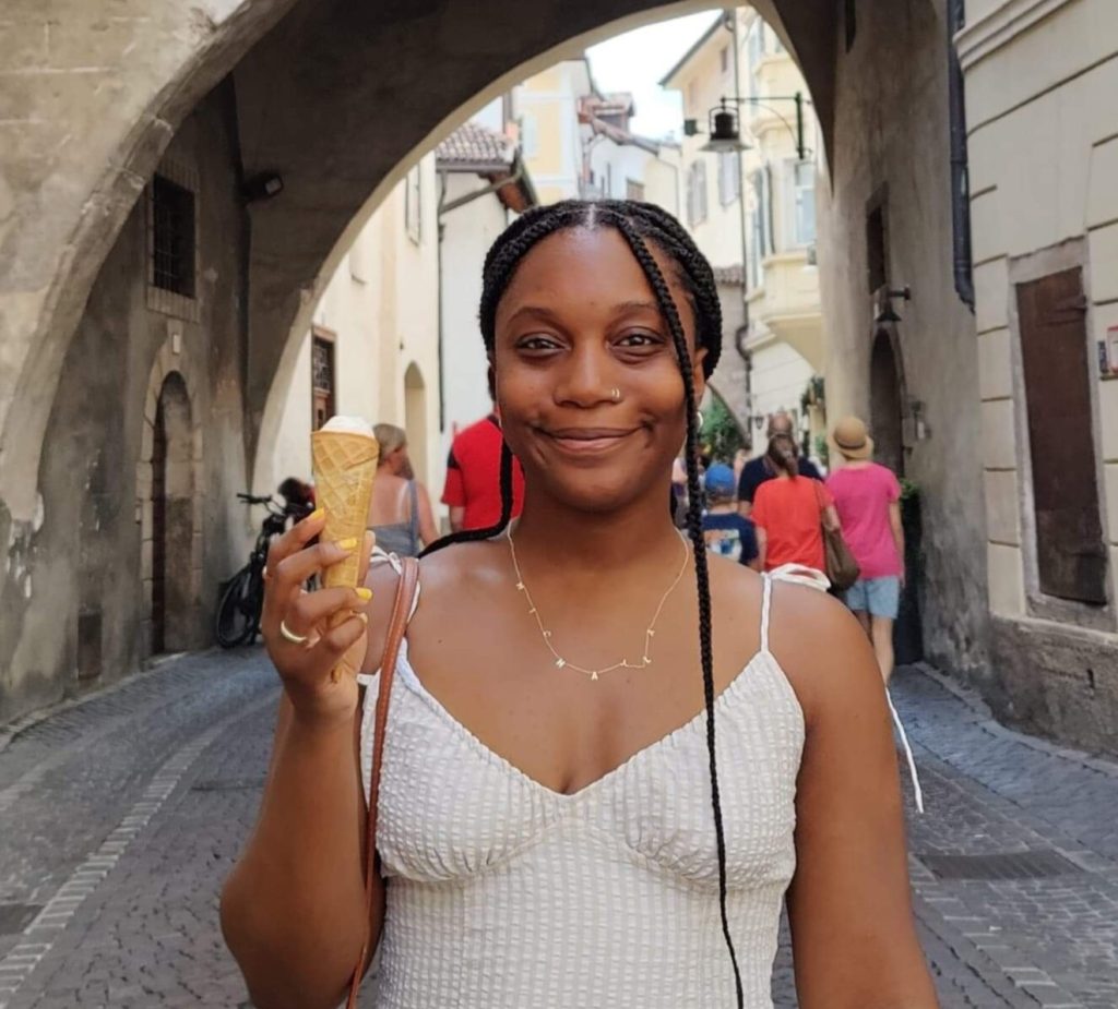 A smiling young black woman with long weaved hair, wearing a white dress. She holds an ice cream cone in her right hand. She is standing in a street with an arch behind her