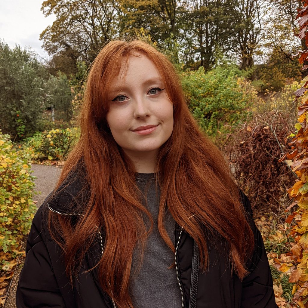 A young white woman with red long hair wearing a black shirt and coat. In the background autumnal trees and shrubs