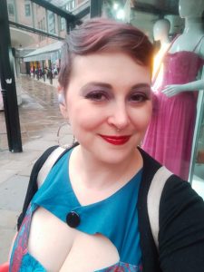 A white woman with short cropped auburn hair, a blue dress, and bright red lipstick gives a small smile to the camera.