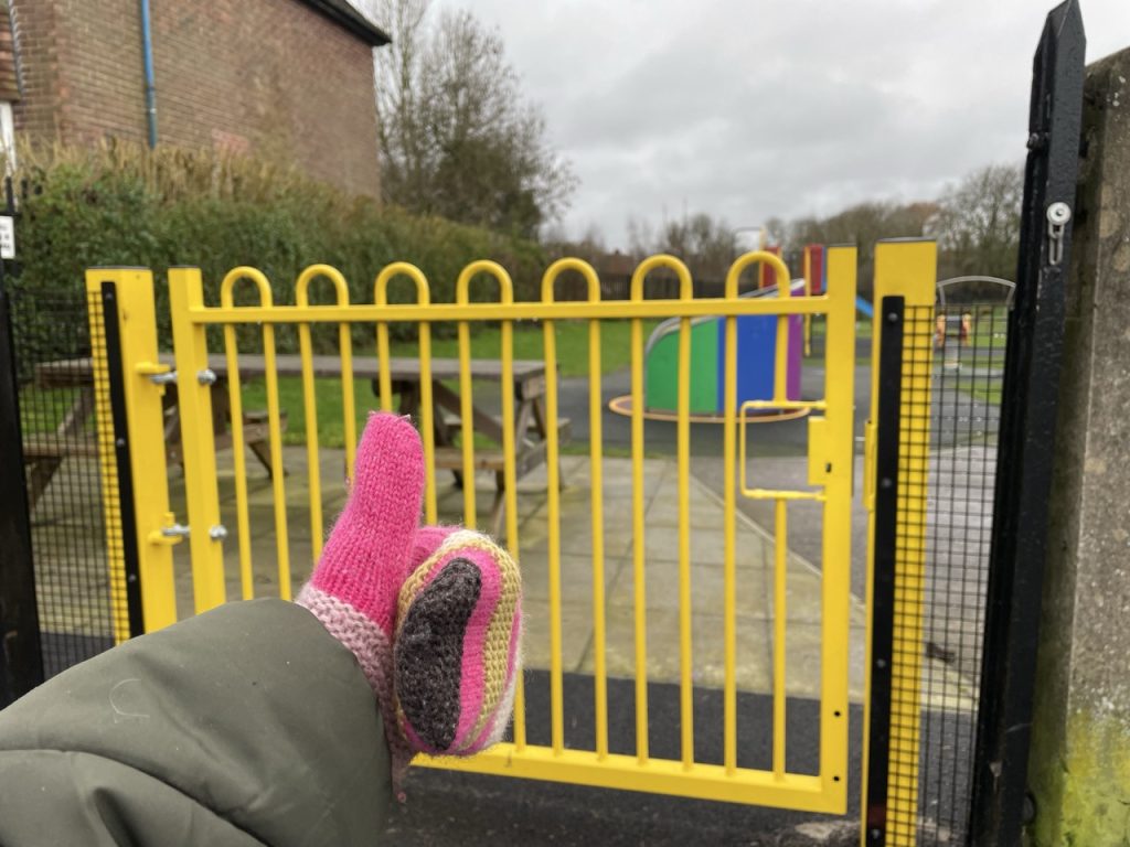 A hand wearing a mitten, giving a thumbs up to a park gate.