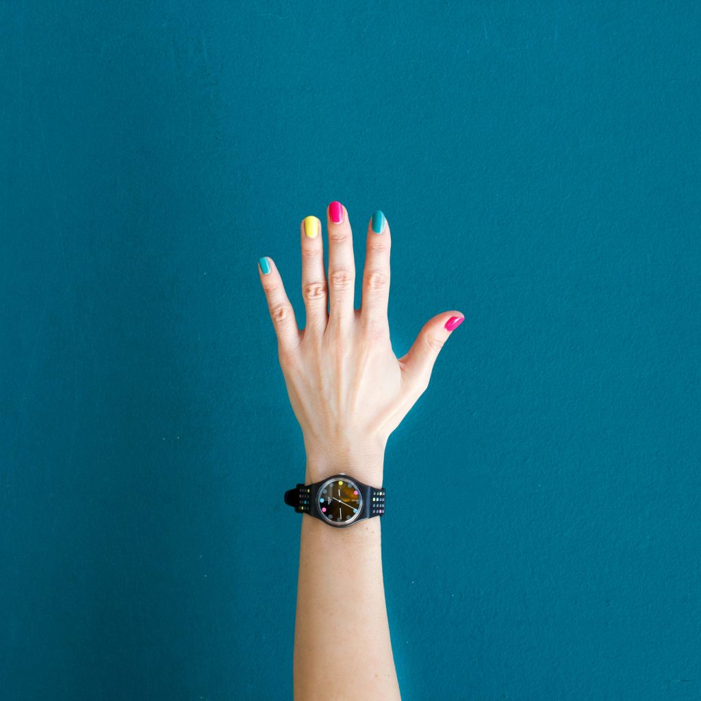 A raised hand with colourfully painted nails, wearing a wristwatch.